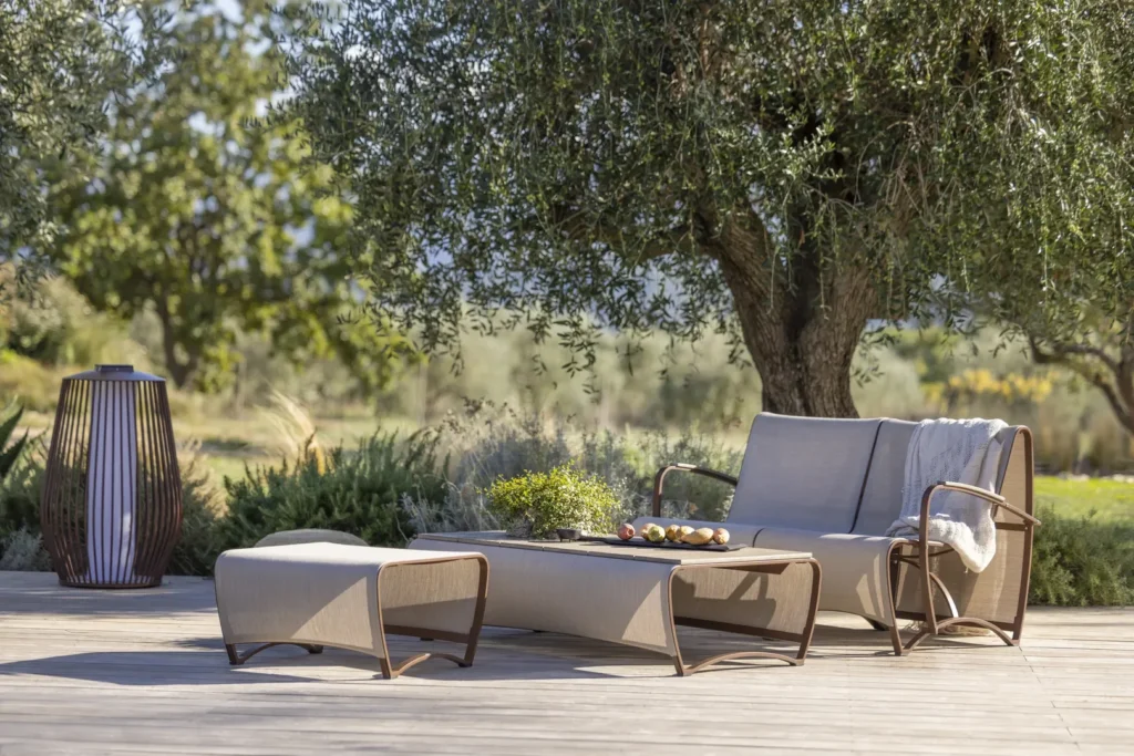 Outdoor patio seating area with modern furniture, a lounge chair, a bench, a coffee table with fruit and plants, and a lantern, set on a wooden deck under a large tree.