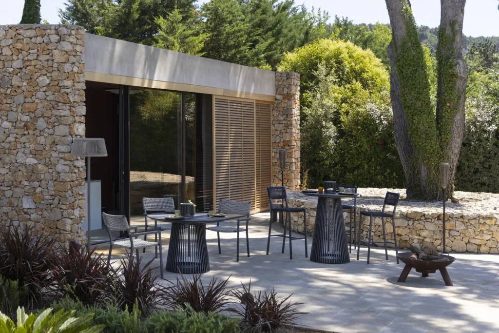 Modern outdoor patio with stone walls, glass doors, black tables and chairs, and surrounding greenery under bright daylight.