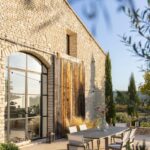 Outdoor dining table with six chairs on a stone patio beside a rustic stone building with large arched windows and wooden doors, surrounded by plants and trees.