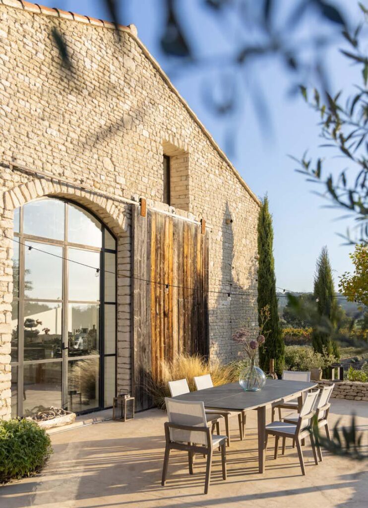 Outdoor dining table with six chairs on a stone patio beside a rustic stone building with large arched windows and wooden doors, surrounded by plants and trees.