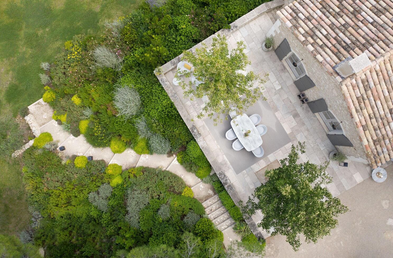 Aerial view of a stone patio with a dining table and chairs, adjacent to a house and surrounded by lush green landscaped garden paths.
