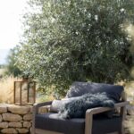 A wooden outdoor armchair with dark cushions and a fluffy pillow sits on a patio next to a stone wall, with a lantern and leafy tree in the background.
