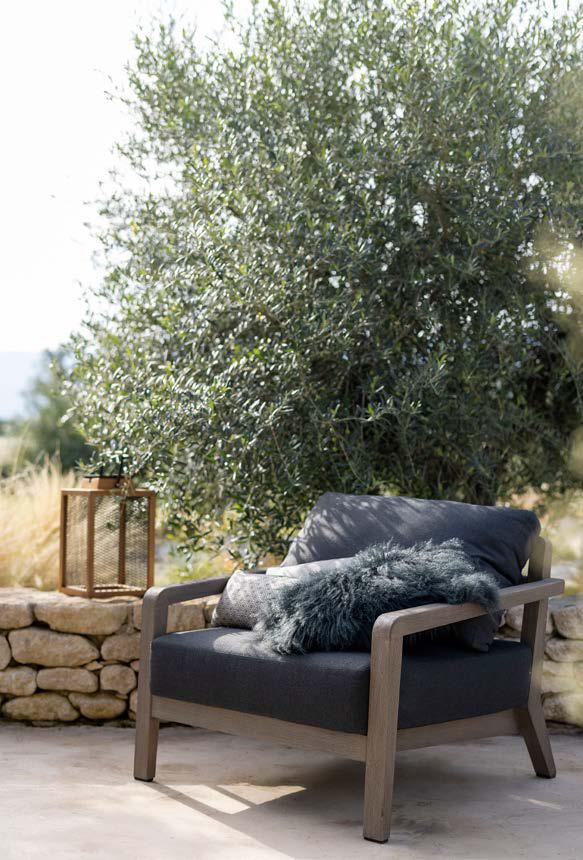 A wooden outdoor armchair with dark cushions and a fluffy pillow sits on a patio next to a stone wall, with a lantern and leafy tree in the background.