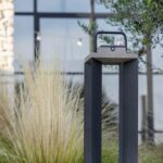 A modern, rectangular outdoor lantern with a handle sits on a stone edge, surrounded by tall grass, with a building and arched window in the background.
