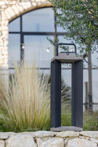 A modern, rectangular outdoor lantern with a handle sits on a stone edge, surrounded by tall grass, with a building and arched window in the background.