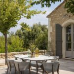 Outdoor patio with a rectangular dining table and six chairs on a stone terrace beside a stone house, surrounded by trees and greenery under a partly cloudy sky.