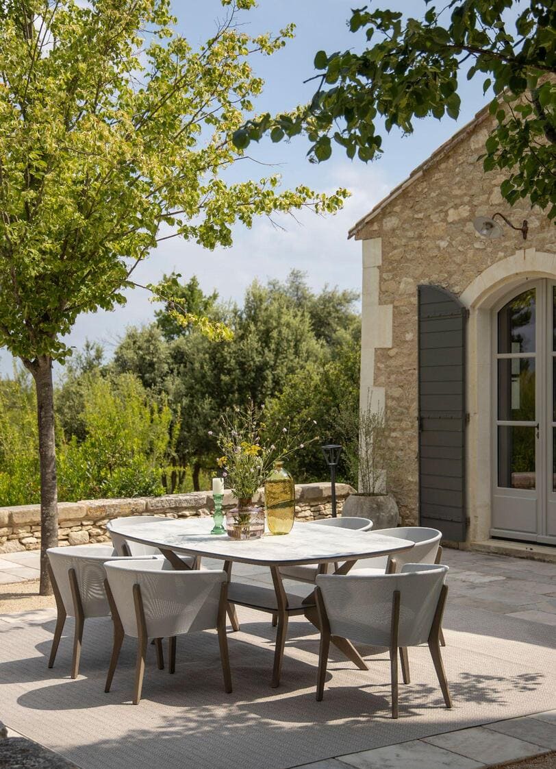 Outdoor patio with a rectangular dining table and six chairs on a stone terrace beside a stone house, surrounded by trees and greenery under a partly cloudy sky.
