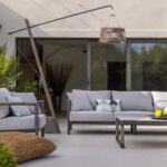 Modern outdoor patio with a light gray sofa set, coffee table with lemons, potted plants, and a woven pendant light, set against sliding glass doors and a beige wall.