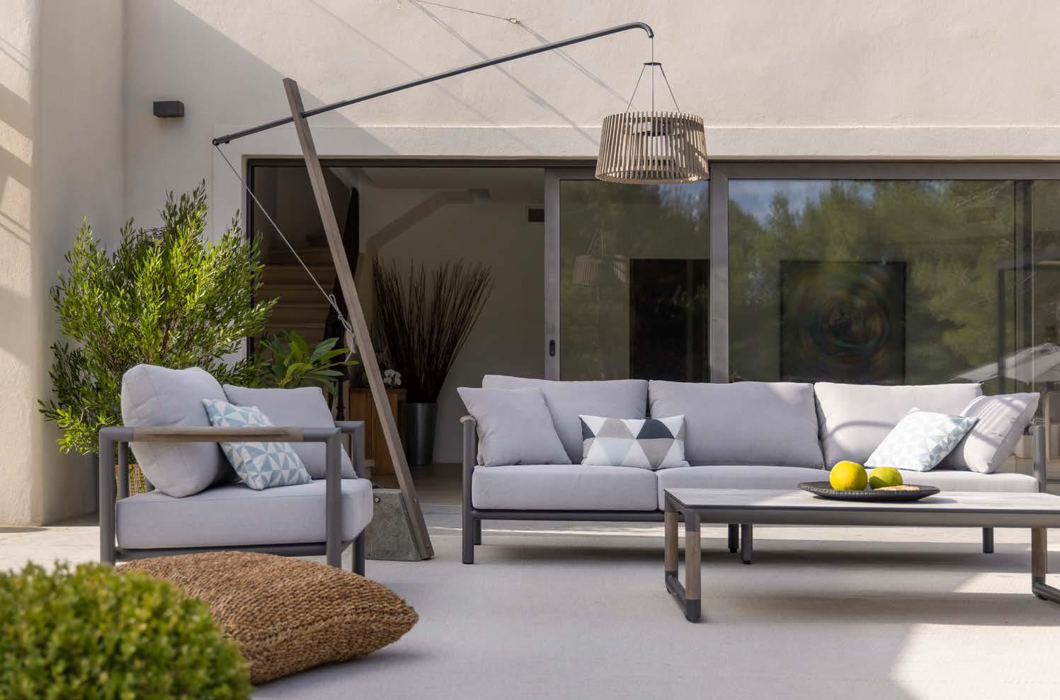 Modern outdoor patio with a light gray sofa set, coffee table with lemons, potted plants, and a woven pendant light, set against sliding glass doors and a beige wall.