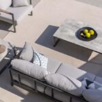 Outdoor patio with gray cushioned furniture, a concrete coffee table holding a black bowl with green apples, and a beige rug on tiled flooring.