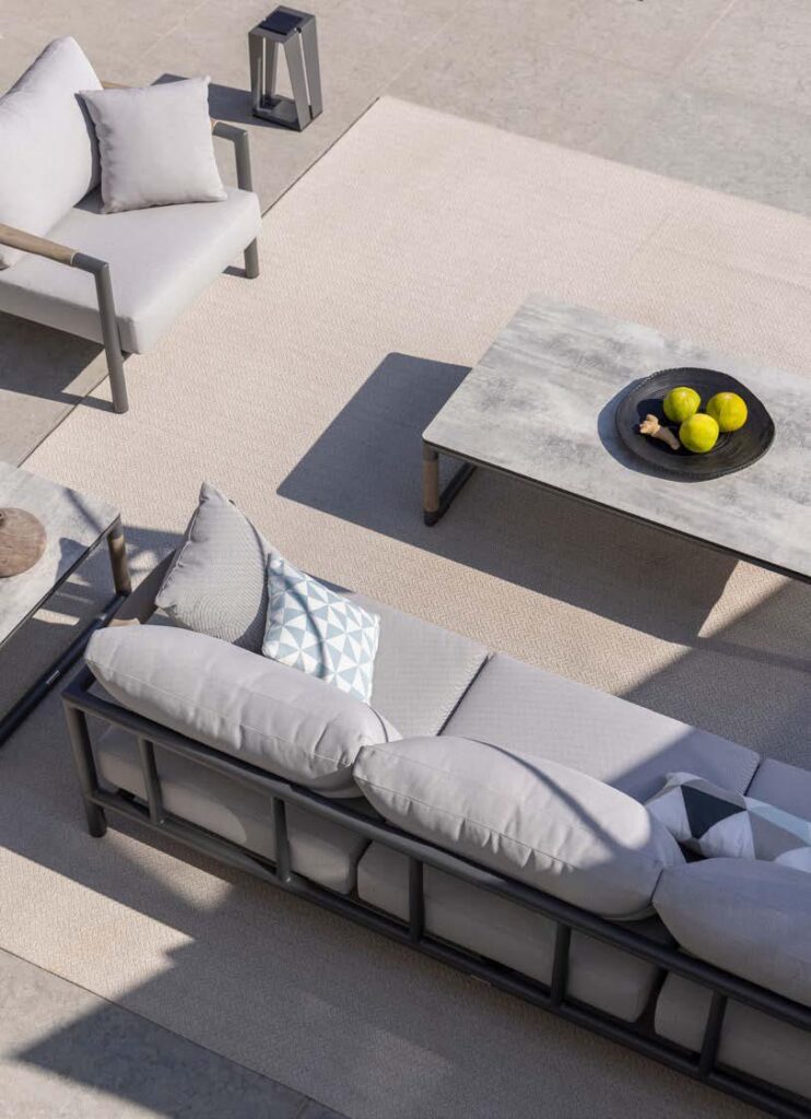 Outdoor patio with gray cushioned furniture, a concrete coffee table holding a black bowl with green apples, and a beige rug on tiled flooring.