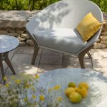 A modern outdoor chair with a yellow cushion sits on a stone patio next to a small table; a glass plate with lemons is on a larger table in the foreground. Greenery is in the background.