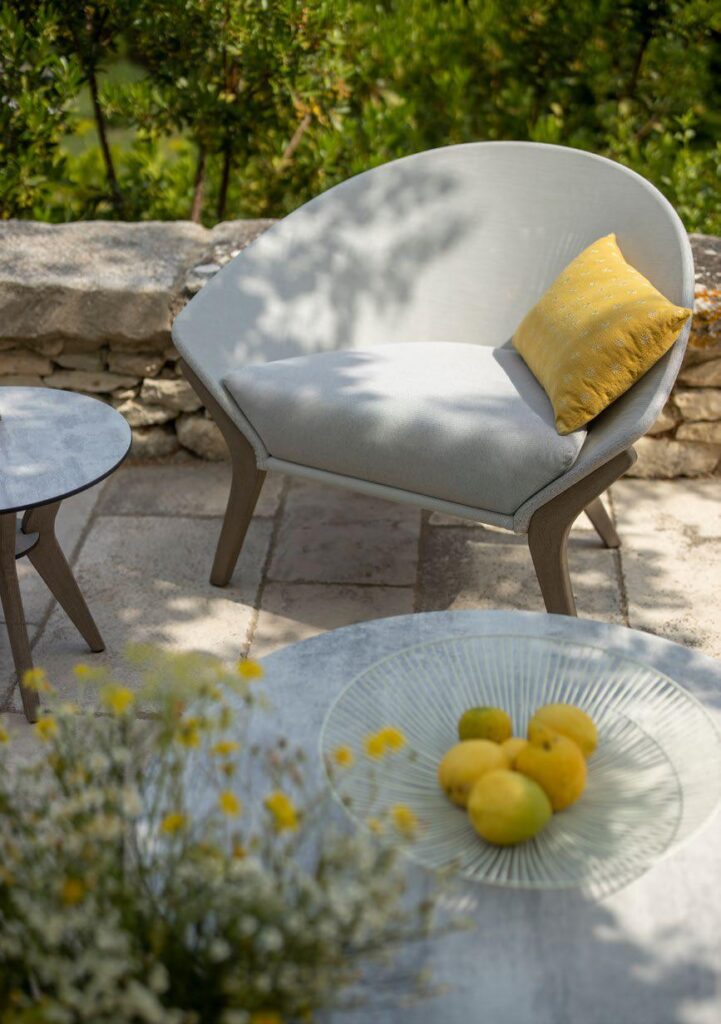 A modern outdoor chair with a yellow cushion sits on a stone patio next to a small table; a glass plate with lemons is on a larger table in the foreground. Greenery is in the background.