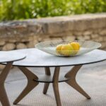 A low, round table with wooden legs holds a decorative bowl filled with yellow lemons, set outdoors near a stone wall and greenery.