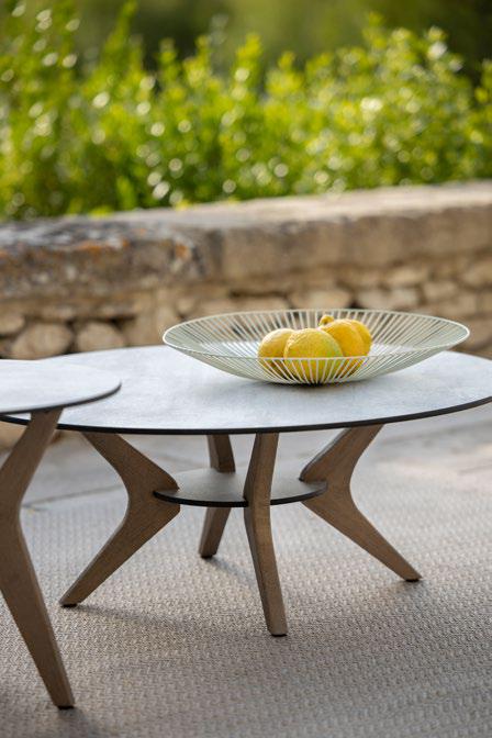 A low, round table with wooden legs holds a decorative bowl filled with yellow lemons, set outdoors near a stone wall and greenery.