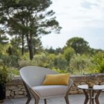 Modern outdoor chair with a yellow pillow sits on a patio next to small round tables, with trees and greenery in the background.