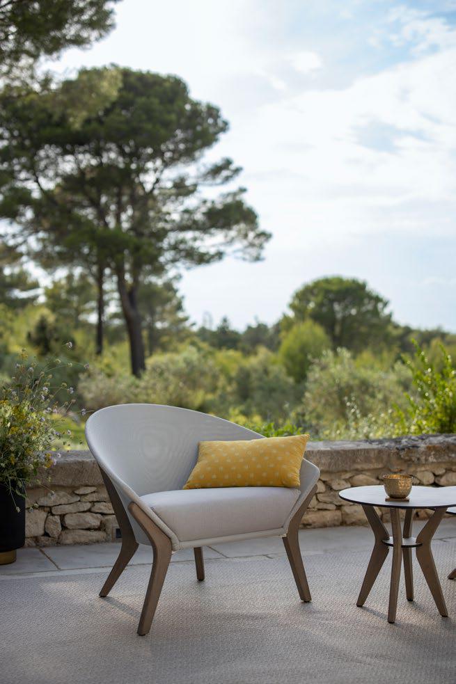 Modern outdoor chair with a yellow pillow sits on a patio next to small round tables, with trees and greenery in the background.