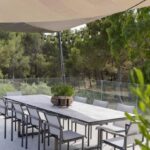 A long outdoor dining table with ten chairs is set under a beige sunshade and string lights, surrounded by glass railing and trees in the background.
