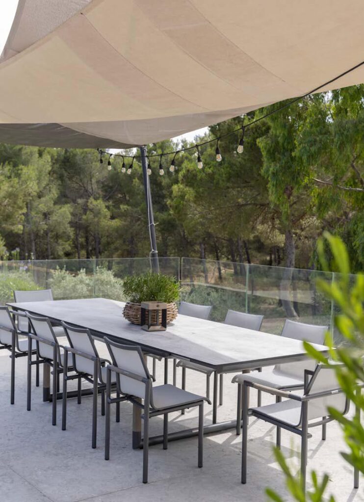A long outdoor dining table with ten chairs is set under a beige sunshade and string lights, surrounded by glass railing and trees in the background.