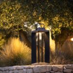 A modern black metal lantern with a handle emits warm light, placed on a stone wall outdoors amid grass and trees at dusk.