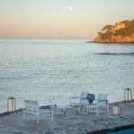 White patio furniture and lanterns are arranged by a pool overlooking a calm sea at sunset, with a cliff and house in the background and the moon visible in the sky.