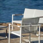 A white metal chair sits on a stone patio near the water, with another chair and a small table nearby.