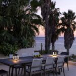 Outdoor dining table with eight chairs and two lanterns on a patio, surrounded by palm trees, overlooking the ocean at sunset.