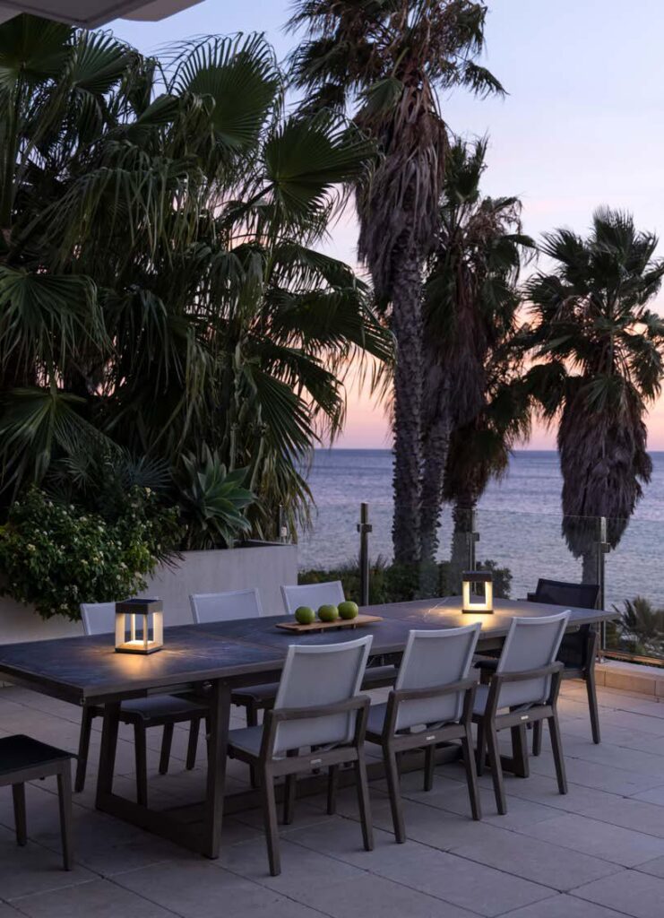 Outdoor dining table with eight chairs and two lanterns on a patio, surrounded by palm trees, overlooking the ocean at sunset.