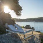 Two white lounge chairs are set on a stone patio overlooking a calm lake, with sunlight streaming through a tree and hills visible in the background.