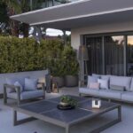 Modern outdoor patio with gray sofas, a large square coffee table holding a plant and lantern, potted plants, and a decorative turtle figure on the ground. Balcony and greenery in background.