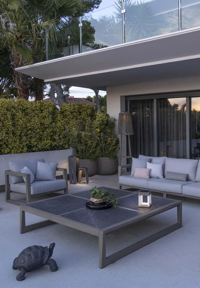 Modern outdoor patio with gray sofas, a large square coffee table holding a plant and lantern, potted plants, and a decorative turtle figure on the ground. Balcony and greenery in background.