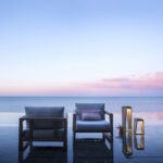 Two modern lounge chairs and two lanterns are placed on a reflective terrace overlooking a calm sea at sunset, with a clear sky and pink clouds.