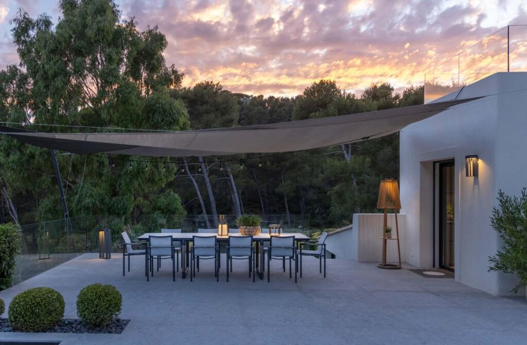 Outdoor dining area with a long table and chairs under a shade sail, next to a modern white building, at sunset with trees in the background.
