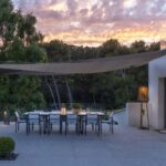Outdoor dining area with a long table and chairs under a shade sail, next to a modern white building, at sunset with trees in the background.