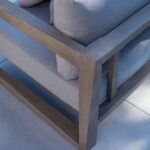 Close-up of a modern outdoor chair with light gray cushions, a wooden frame, and a patterned pillow on a tiled surface.