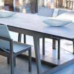 A modern wooden dining table with two white chairs and two decorative shallow bowls, set in a bright room with large windows and stone walls.