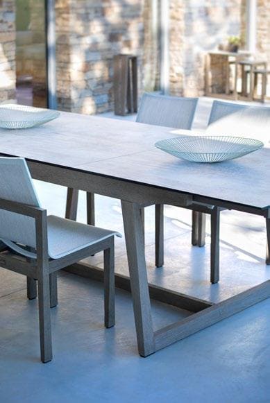 A modern wooden dining table with two white chairs and two decorative shallow bowls, set in a bright room with large windows and stone walls.