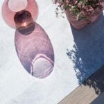 A pink glass vase casts a purple shadow on a light surface next to a potted plant with small pink flowers.