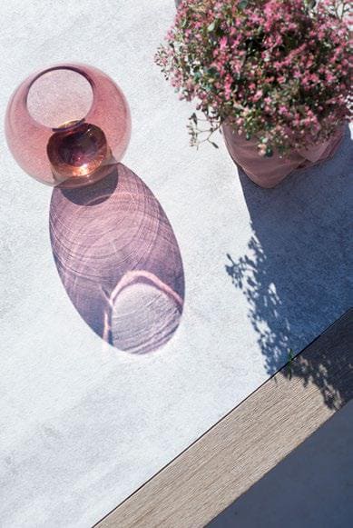 A pink glass vase casts a purple shadow on a light surface next to a potted plant with small pink flowers.