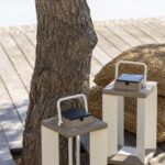 Two rectangular lanterns with wooden tops and white frames are placed on a wooden deck next to a tree and a woven cushion, with a pool in the background.