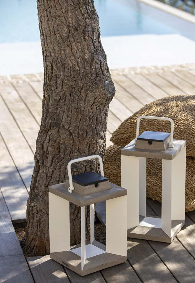 Two rectangular lanterns with wooden tops and white frames are placed on a wooden deck next to a tree and a woven cushion, with a pool in the background.