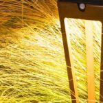 A modern outdoor lamp illuminates tall yellow grass at night, casting a bright, warm glow over the surrounding vegetation.