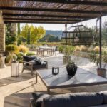 A covered patio with lounge chairs, a table, potted plants, and a view of an outdoor dining area surrounded by greenery and trees.