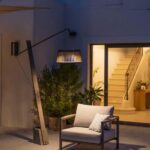 A cushioned armchair sits outdoors under a modern floor lamp near a building entrance with an open door and visible staircase.