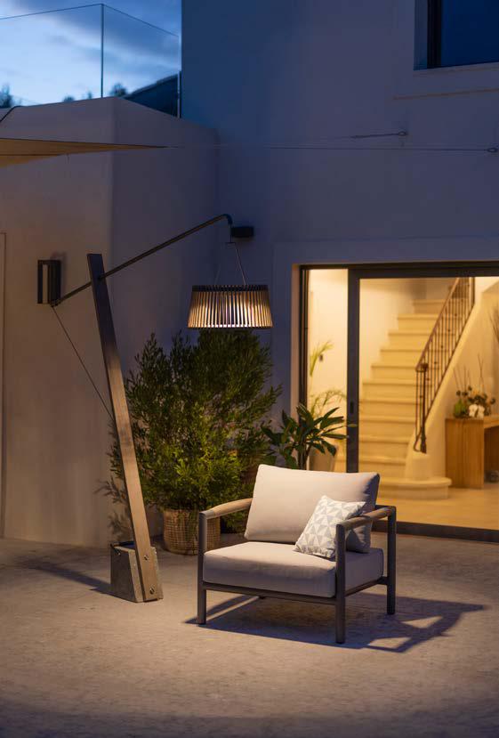 A cushioned armchair sits outdoors under a modern floor lamp near a building entrance with an open door and visible staircase.
